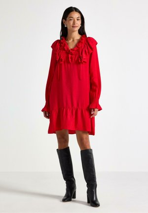 Rød langærmet kjole med flæser ved halsudskæringen og bunden, der har en løstsiddende pasform og en struktureret stof. Bæres med knæhøje sorte støvler.