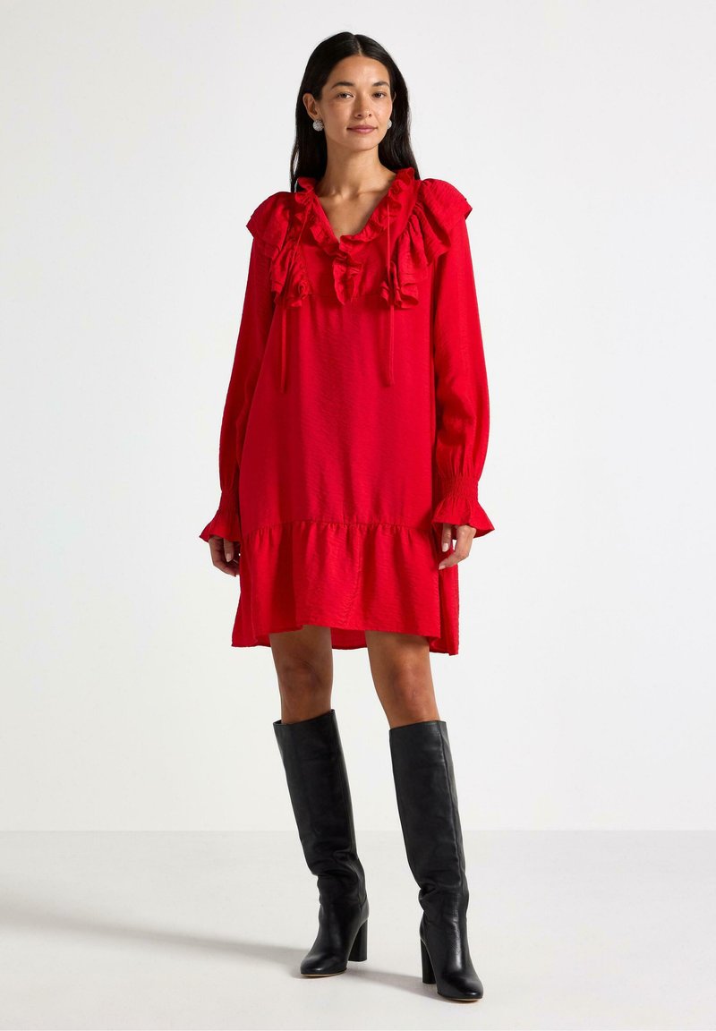 Rød langærmet kjole med flæser ved halsudskæringen og bunden, der har en løstsiddende pasform og en struktureret stof. Bæres med knæhøje sorte støvler.