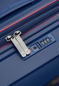 Valigetta rigida di colore blu navy con superficie texture, dettagli in zip rossi, doppie zip metalliche argento e un meccanismo di chiusura approvato TSA.