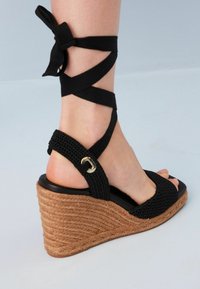 Sandalo wedge intrecciato nero con lacci alla caviglia, caratterizzato da una piattaforma in juta testurizzata e un accento dorato sulla fascia. Design open toe.