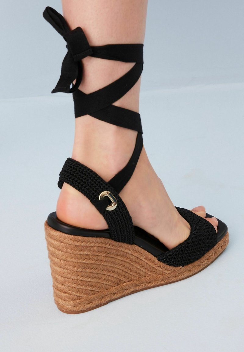 Sandalo wedge intrecciato nero con lacci alla caviglia, caratterizzato da una piattaforma in juta testurizzata e un accento dorato sulla fascia. Design open toe.