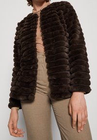 Brun faux fur-jacka med horisontella ränder och en bekväm textur; har en rund krage och öppen framside-design, i kombination med ljusbeige byxor.