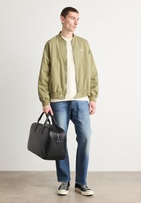 Groene bomberjack, crème trui met ronde hals, blauwe jeans, zwarte leren reistas en zwarte sneakers met witte zolen. Eenvoudige, casual outfit.