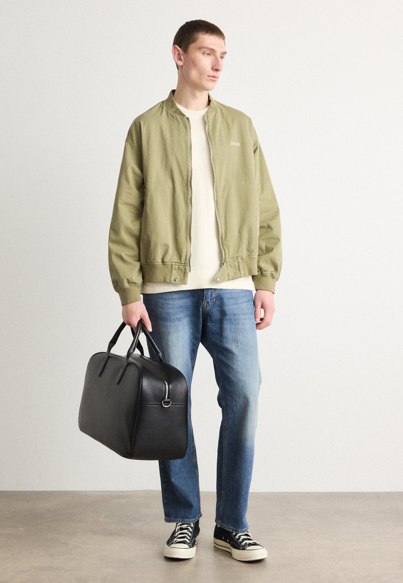 Grüne Bomberjacke, cremefarbener Rundhalsausschnitt-Pullover, blaue Jeans, schwarze Leder-Duffle-Tasche und schwarze Sneakers mit weißen Sohlen. Einfaches, lässiges Outfit.