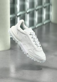 Baskets athlétiques blanches avec tige en cuir, semelle en caoutchouc texturée et accents en mesh contrastants. Présente un logo sur le côté et une fermeture à lacets.