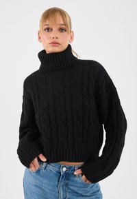 Maglione a collo alto in maglia nera con design cropped, polsini a coste e collo a imbuto spesso, abbinato a jeans blu a vita alta.