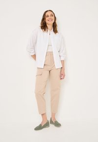 Witte overhemd met knopen, beige cargobroek en groene instapschoenen. Het shirt heeft een zak, en de broek heeft zijzakken en een taps toelopende pasvorm.