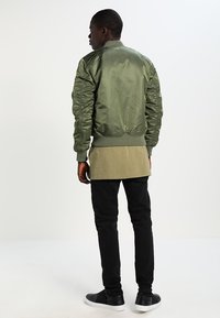 Olive green bomber jacket with ribbed cuffs and hem, featuring a smooth texture and a relaxed fit, paired with black pants and sneakers.