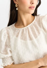 Blusa bianca con maniche a palloncino, decorata con piccole macchie in rilievo. Presenta un collo arricciato e polsini elasticizzati. Orecchini dorati con dettagli in perla.