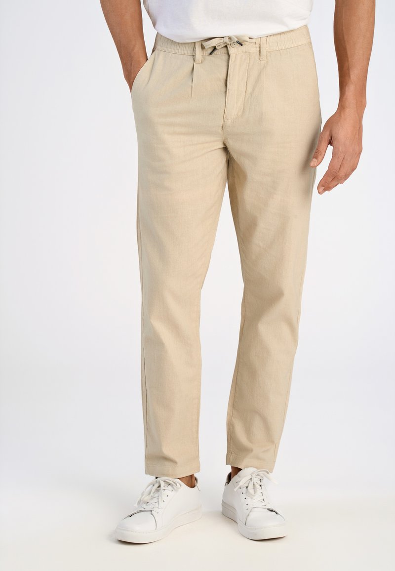 Beige lichte broeken met zijzakken, voorzien van een elastische tailleband en een trekkoordsluiting, gecombineerd met witte sneakers.