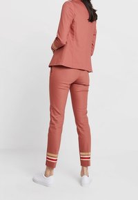 Formsydd rosafärgad kavaj och matchande byxor med guld- och rödrandiga accenter vid ankeln. Tillverkad av mjukt, elastiskt material. Vita sneakers.