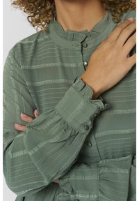 Blusa verde chiaro, texturizzata, con strisce verticali. Collo e polsini arricciati, chiusure con bottoni e dettagli con maniche ampie.