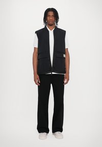 Black sleeveless vest with textured edges, front pockets, worn over a white polo shirt and paired with black wide-leg pants. White sneakers complete the outfit.