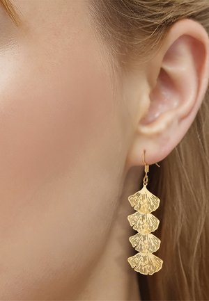 Nahaufnahme eines Frauenohrs mit einem goldenen Anhängerohrring, der aus vier fächerförmigen, strukturierten Blattsegmenten besteht.