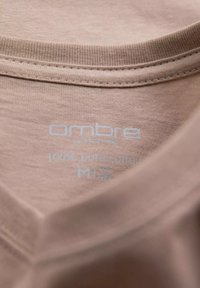 Světle béžový bavlněný T-shirt s límečkem, na kterém je štítek s textem "Ombre 100% čistá bavlna" ve světle modré barvě, s měkkou a hladkou texturou.