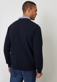 Navy gebreide cardigan met een textuurpatroon, geribde boorden en zoom, gedragen over een lichtblauw overhemd met kraag, gecombineerd met blauwe spijkerbroek.