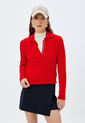Czerwony sweter w splot warkoczowy z głębokim dekoltem w kształcie V i ściągaczami przy mankietach. Połączony z granatową spódnicą typu wrap i jasnoszarym czapka. Gładki, teksturowany materiał.