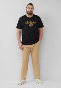 Schwarzes Baumwoll-T-Shirt mit dem Aufdruck "s.Oliver Elevated Essentials" in Gelb. Kombiniert mit beigen Hosen und hellen Turnschuhen. Schlichtes Design.