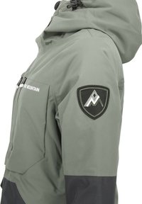 Veste étanche en vert olive avec des accents noirs. Présente une capuche, des poches zippées et un patch logo en forme de bouclier sur la manche gauche.