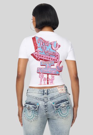 Vrouw met kort zwart haar die een wit T-shirt draagt met een kleurrijke vintage bordgrafiek en vervaagde blauwe spijkerbroek met gedetailleerde achterzakken.