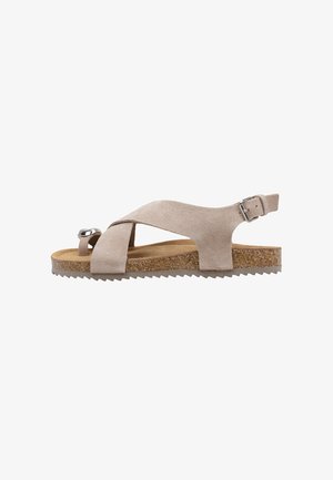 Sandalia de ante beige con tiras cruzadas, correa ajustable con hebilla en el talón, suela de corcho y anillo en el dedo con detalle metálico, plana con suela exterior estriada.