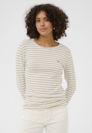 Femme aux cheveux bouclés portant un chemisier à manches longues beige et blanc à rayures avec un petit cœur rose sur le côté gauche de la poitrine et un pantalon blanc.