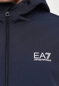 Tengerészeti kék kapucnis dzseki fekete cipzárral és fehér EA7 Emporio Armani logóval a mellkason.