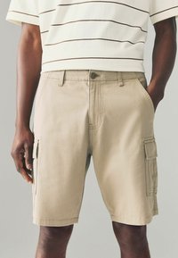 Beige cargo shorts valmistettu puuvillasta, jossa on sivutaskut ja etupainonappi. Suunnittelussa on kontrastiväriset ompelu yksityiskohdat.