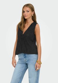 Zwarte mouwloze top met een V-hals, versierd met geborduurde bloemenpatronen en een losse pasvorm, gecombineerd met lichtblauwe denim jeans.