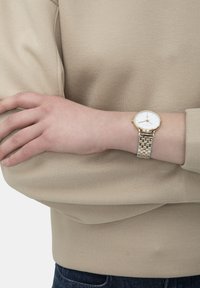 Silber- und goldfarbene Uhr mit einem runden weißen Ziffernblatt, im Armband-Stil aus Metall, an einem Handgelenk getragen, das ein beiges Langarmoberteil trägt.