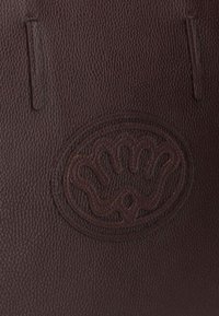 Bruine leren textuur met een centraal verhoogd cirkelvormig logo, voorzien van een ingewikkeld handontwerp en eenvoudige uitsparingen.