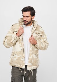 Beige Jacke mit Tarnmuster, Kapuze, Druckknopfverschluss und mehreren Taschen; mit verstellbarem Kordelzug am Saum.