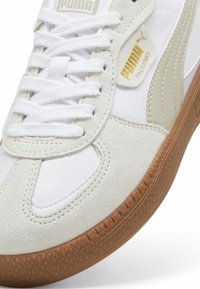 Baskets blanches avec des accents beige, comprenant une tige en daim texturé et en cuir lisse, des lacets plats et une semelle extérieure en caoutchouc naturel. Détail du logo doré.