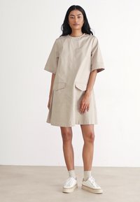 Vestido bege de manga curta com decote redondo, dois bolsos frontais e um corte descontraído, combinado com ténis brancos e meias claras.