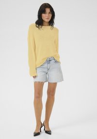Kollane kudumissweater, millel on avar lõige, koos helesiniste denimsärkidega, mille ääred on kulunud, ja mustade terava ninalt kontsadega.