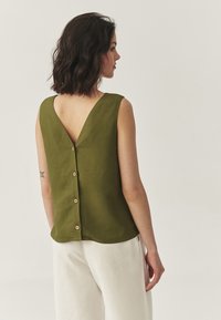 Bluză fără mâneci de culoare verde măsliniu, cu un decolteu adânc la spate, având șapte nasturi din lemn de-a lungul coloanei vertebrale. Material moale, texturat.