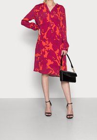 Vestito a maniche lunghe rosso e rosa con motivo, scollo a V e chiusura con bottoni. Abbinato a una borsa nera e tacchi neri con cinturini.