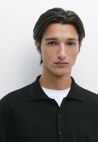 Schwarzes Polohemd mit kurzem, kragenförmigem Ausschnitt und geknöpften Knopfleisten, über einem weißen T-Shirt getragen. Glatte Textur, minimalistisches Design.