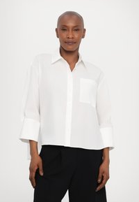 Hvit skjorte med knapper laget av lett materiale, med krage, lange ermer og en enkelt brystlomme. Enkel og minimalistisk design.