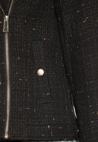 Czarna kurtka o fakturowanej powierzchni z srebrnym zamkiem błyskawicznym, z złotymi nitkami, przednia kieszeń na zatrzask oraz dopasowany krój.