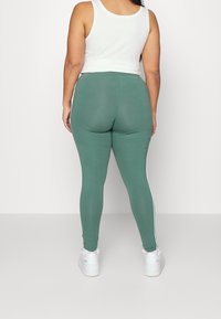 Gröna leggings med en slät textur, vita sidostrecken, hög midja och en figurnära design. Matchas med vita sportskor och en kort vit tanktop.