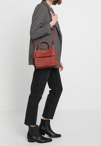 Sac à main en cuir rouge avec un design structuré, des poignées supérieures et une bandoulière réglable, associé à un blazer à carreaux et un jean noir.