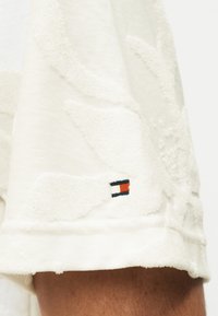 Λευκό υφασμάτινο σόρτς με διακριτικό φυλλώδες σχέδιο και μικρό ορθογώνιο λογότυπο σε κόκκινο, λευκό και μαύρο κοντά στη μανσέτα, πάνω στο πόδι ενός ατόμου.