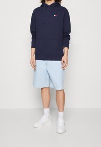 Navy blue hoodie with front pocket and drawstring, paired with light blue denim shorts and white sneakers. Simple design with minimal branding.