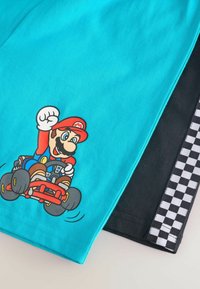 Shorts de algodón color turquesa con un personaje de dibujos animados conduciendo un kart rojo. Tienen un detalle de cuadros en blanco y negro en el lateral.