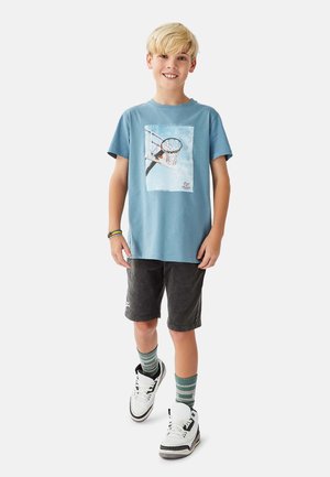 Lyseblå bomuldst-shirt med et basketballmotiv. Båret med mørke shorts, hvide sneakers og sokker med grønne striber.