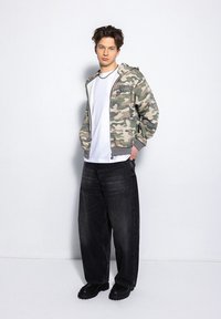 Hoodie w kamuflażu z zamkiem z przodu, szarymi akcentami i logo; w zestawieniu z luźnymi czarnymi dżinsami i czarnymi butami. Neutralne tło.