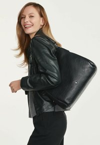 Czarna skórzana torba tote z teksturowaną powierzchnią, prostokątnym kształtem i srebrnymi akcentami, noszona na ramieniu przez modelkę w czarnej kurtce.