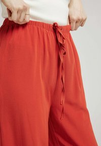 Pantalons légers rouges en tissu avec une taille élastique. Présente un détail de lien à l'avant et une texture lisse. Coupe décontractée et évasée.