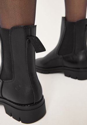 Bottines en cuir noir à semelles épaisses portées avec des collants noirs transparents sur un fond clair.